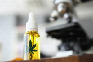 Focus on glass flask with cannabinoid oil and green ganja symbol on it. Laboratory interior with microscope on blurred b