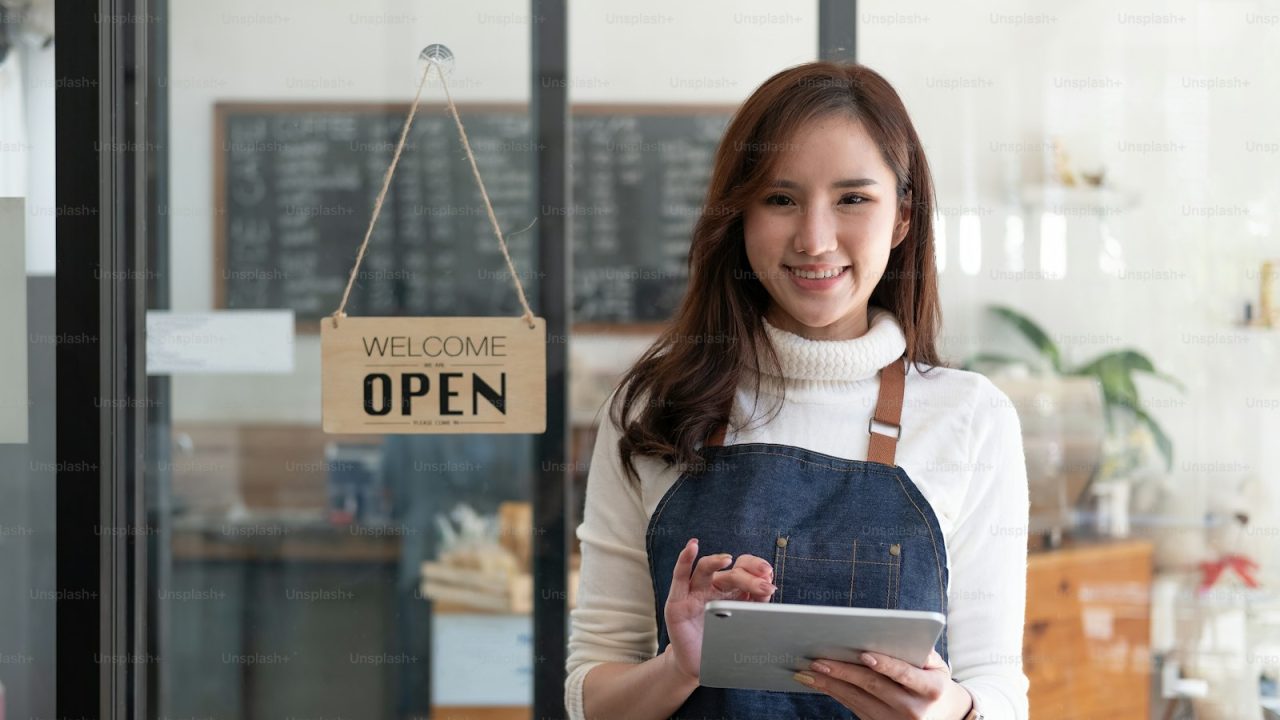 Portrait of a smiling Asian entrepreneur standing behind her cafe counter with open sign board.