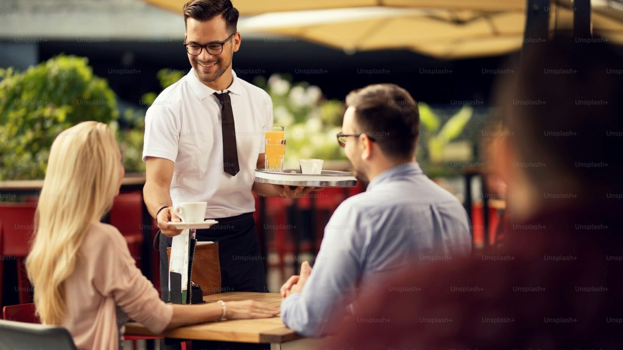 Happy waiter giving coffee to a couple while serving them in an outdoor cafe.