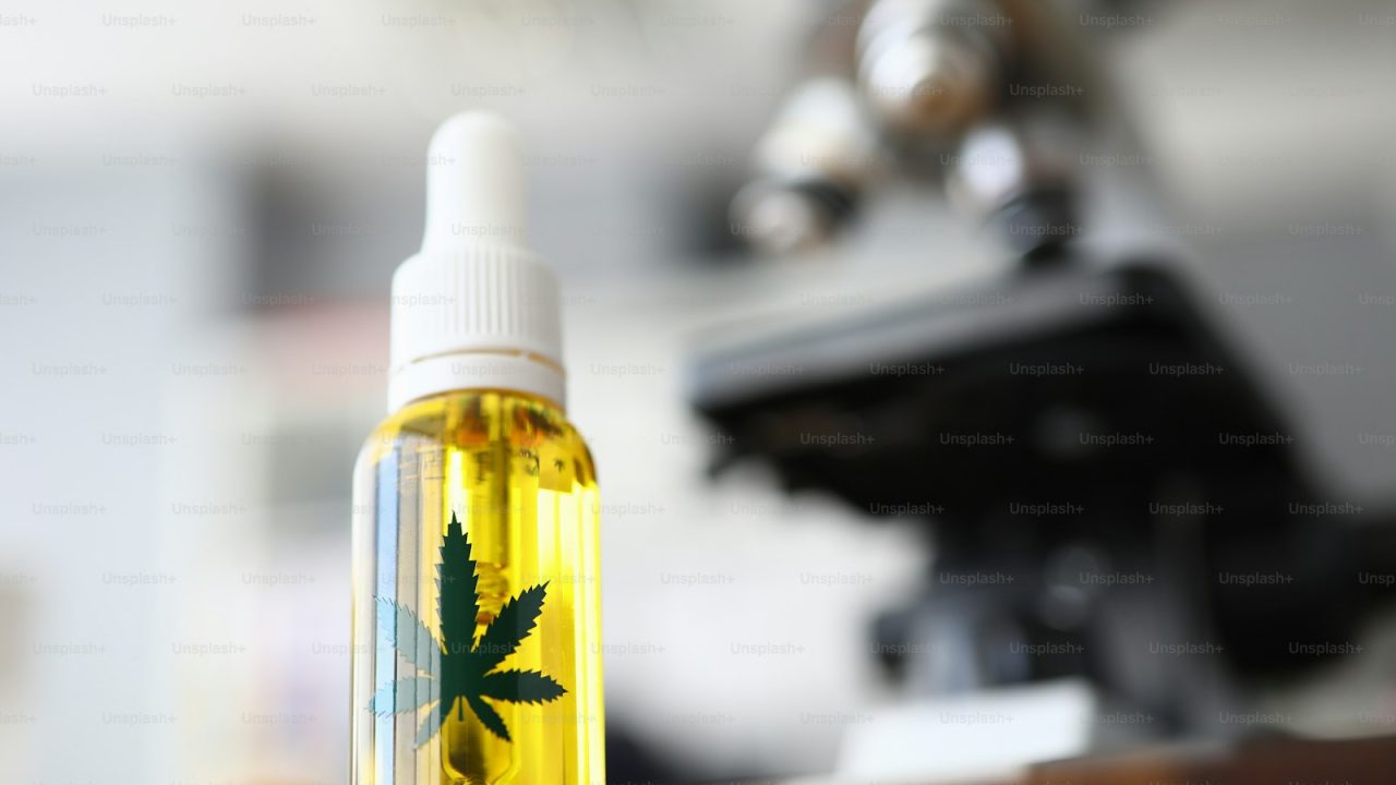 Focus on glass flask with cannabinoid oil and green ganja symbol on it. Laboratory interior with microscope on blurred b