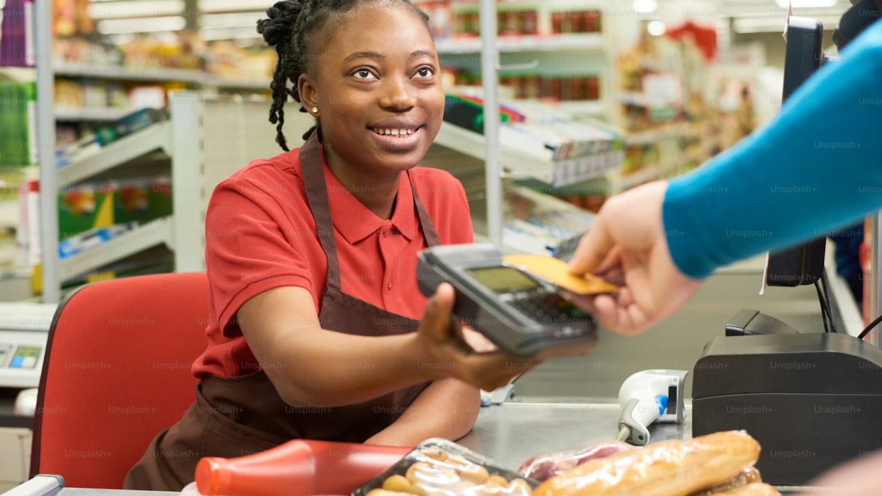 Young smiling salesperson looking at consumer holding credit card over payment terminal while buying food products in su